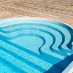 New modern fiberglass plastic swimming pool entrance step with clean fresh refreshing blue water on bright hot summer day at yard or resort hotel spa area. Wooden flooring deck of teak or larch board.