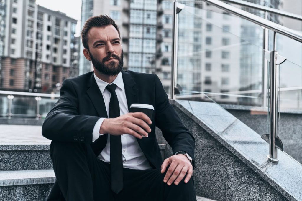Hard decision to make. Good looking young man in full suit holding disposable cup and looking away while sitting on the stairs outdoors