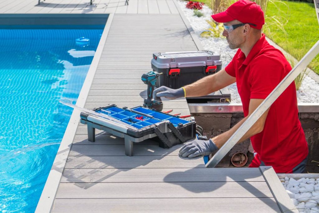 Outdoor Swimming Pool Heating and Filtering System Maintenance Performed by Caucasian Middle Aged Technician. Open Toolbox and Electric Drill Driver on the Floor in Front of Him.
