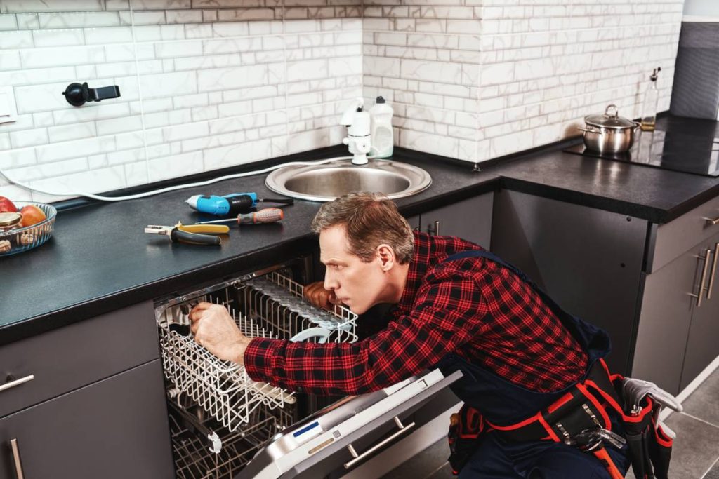 Side view of repairing dishwasher. Male technician sitting near dishwasher with screwdriver in kitchen with instruments