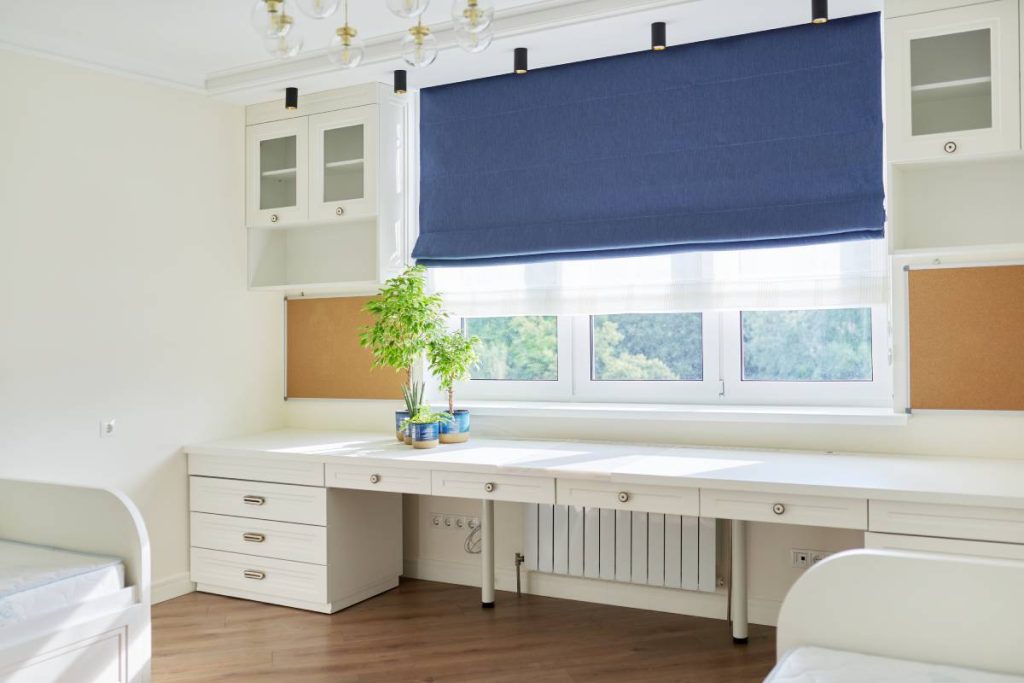 Interior of children's room for two children in light colors. White furniture, table, blue roman blind on the window