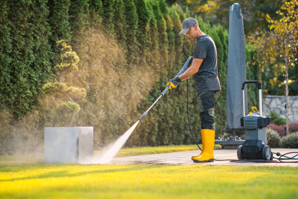 A worker dressed in yellow boots and gloves cleans a patio with a power washer, surrounded by lush greenery on a bright day.