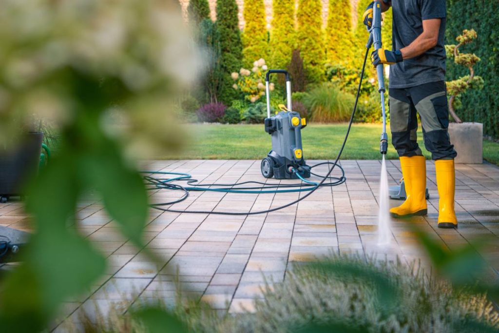 A worker in yellow boots is using a pressure washer to clean patio tiles, surrounded by lush greenery in a beautiful garden.