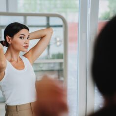 Bored young pretty Asian woman in white t-shirt, beige pants standing at mirror, tired. Korean girl