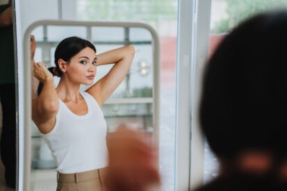 Bored young pretty Asian woman in white t-shirt, beige pants standing at mirror, tired. Korean girl