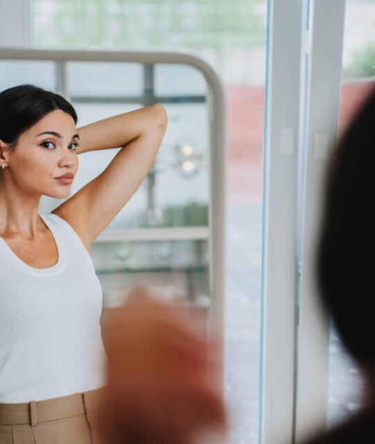 Bored young pretty Asian woman in white t-shirt, beige pants standing at mirror, tired. Korean girl
